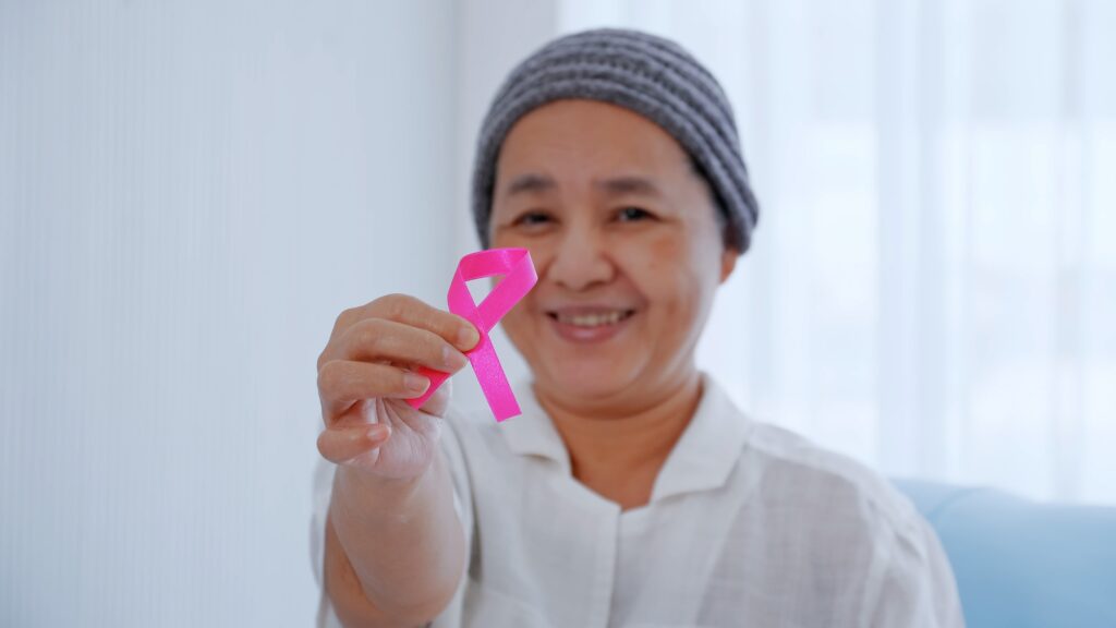 Elderly female patient holding pink ribbon in hospital. Breast cancer awareness concept.